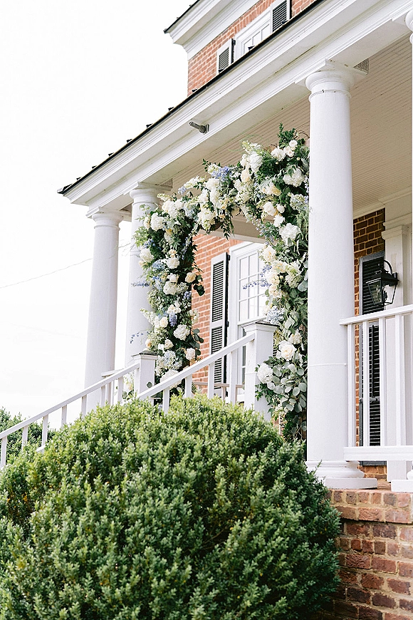 Wedding floral arch with white and blue blooms and greenery on a brick house front porch, framing the steps with eucalyptus accents