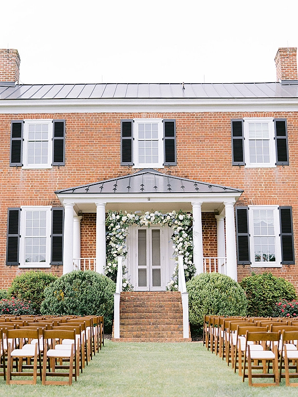 Ceremony setup with outdoor wedding ceremony seating, wood folding chairs facing a white rose and greenery arch at a brick porch entrance