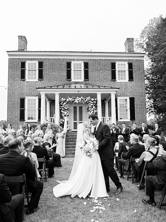Ceremony kiss moment as bride and groom kiss beneath a floral arch, veil flowing, guests lining rose-petal aisle at brick manor porch