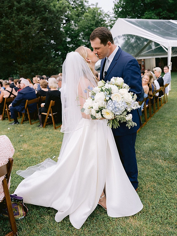 Wedding kiss portrait of newlyweds kissing as the bride holds a rose and hydrangea bouquet, bridal veil flowing by clear tent seating