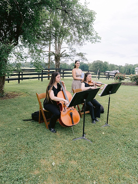 Wedding ceremony musicians, a string duo with cello and violin reading sheet music on stands beside folding chairs on a grassy lawn