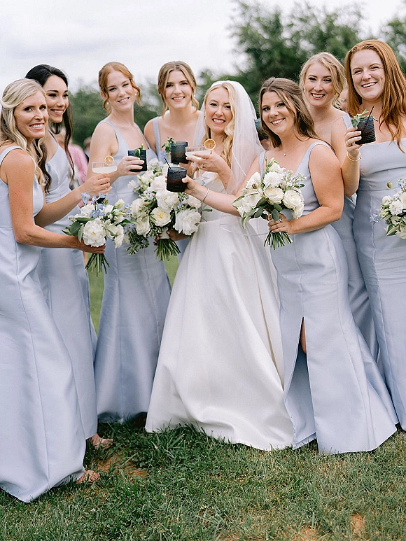Bridesmaid group photo with bride with bridesmaids in light blue dresses, holding white and greenery bouquets and cocktails on a lawn under cloudy sky
