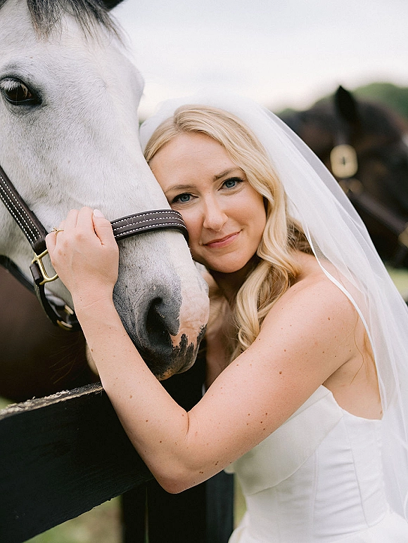 Bridal portrait of a bride with horse, wearing a strapless dress and veil, holding the halter by a wooden fence in a pasture