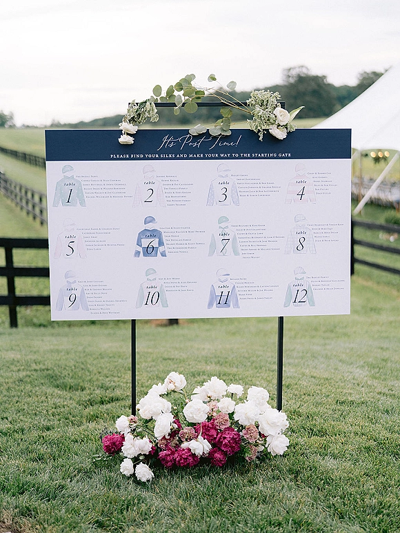 Seating chart sign with wedding seating chart cards in calligraphy on a metal stand, framed by greenery garland and white flowers outdoors
