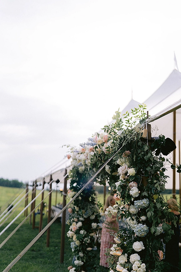 Tent wedding entrance with floral tent entrance garland of roses and hydrangea on wooden posts, rope walkway under canopy lights in open field