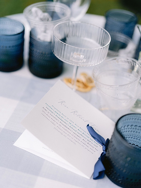 Wedding invitation suite with navy wedding invitation tied in ribbon beside coupe glasses, blue votives, and linen napkin on white tablecloth