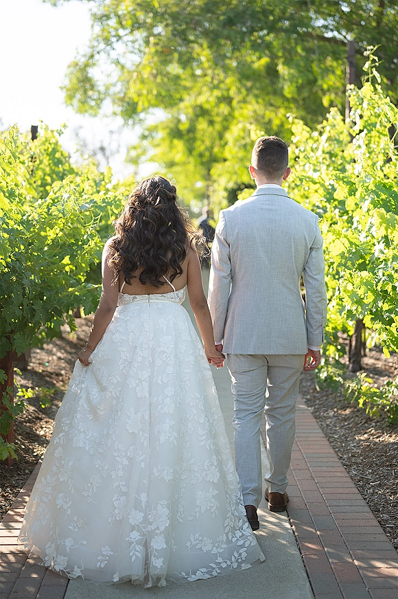 Couple portrait of bride and groom walking away, holding hands down a sunlit vineyard path, lace gown and light gray suit amid greenery
