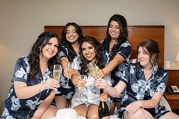 Bridesmaids getting ready in matching satin pajamas raise champagne flutes for a toast on a hotel bed with curled hair and jewelry