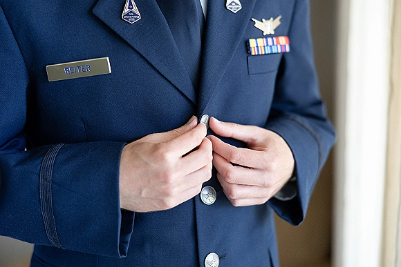 Groom getting ready, groom buttoning jacket on a military dress uniform with service ribbons and lapel insignia in soft window light indoors