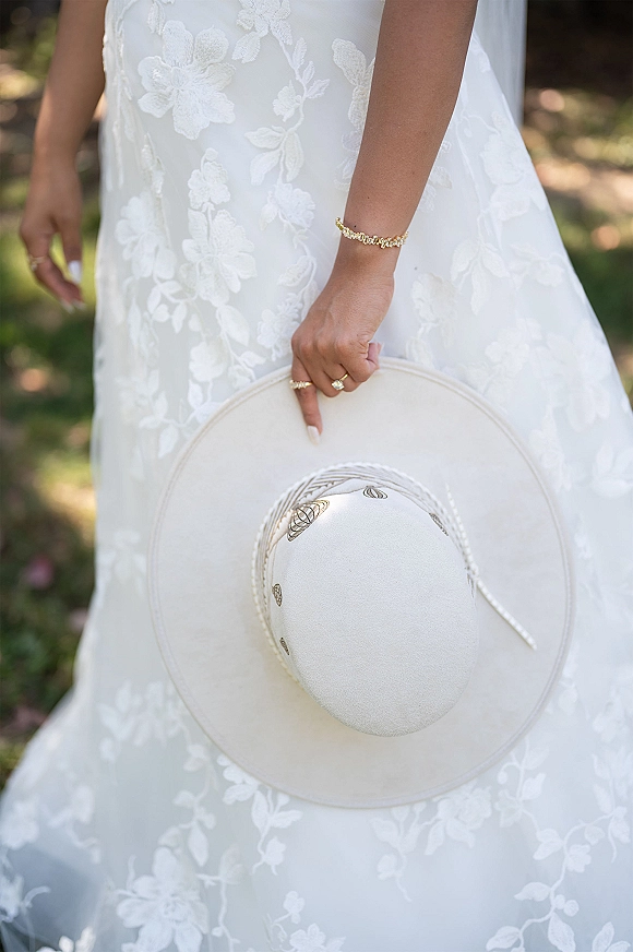 Wedding dress close-up showing lace wedding dress detail as bride holds a wide-brim hat, rings and bracelet visible in sunlit garden greenery