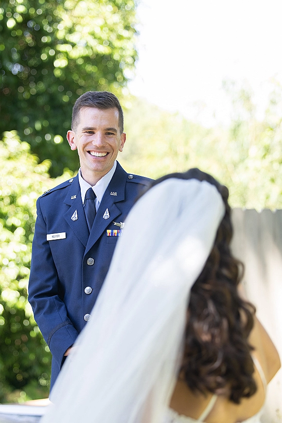 First look moment as bride in veil approaches groom in military dress uniform near green trees and a wooden fence in daylight