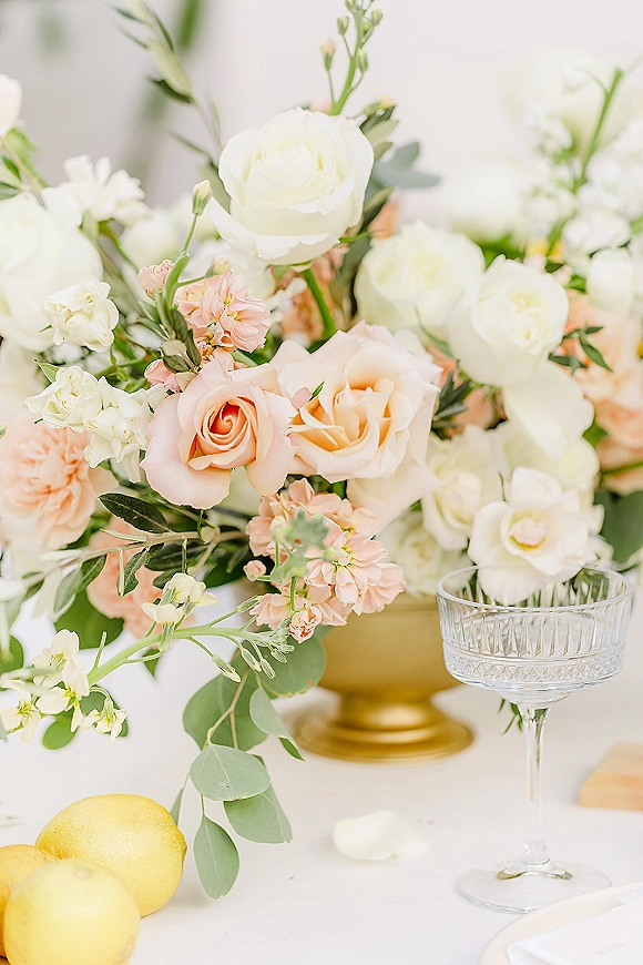 Wedding floral centerpiece in a gold compote centerpiece with white and peach roses, eucalyptus greenery, lemons, and coupe glass on white linen