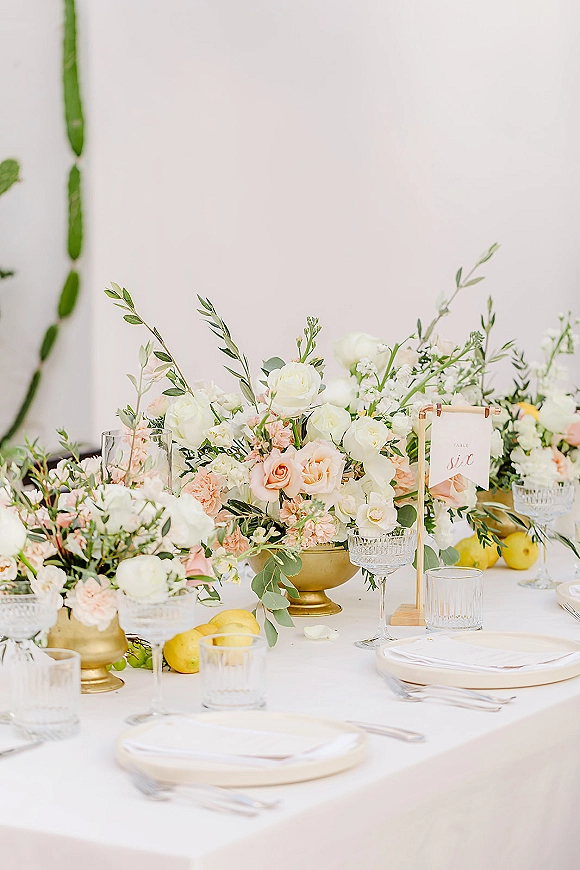 Reception tablescape with wedding table centerpiece of white and blush roses, greenery and lemons in gold compote bowls on white linen