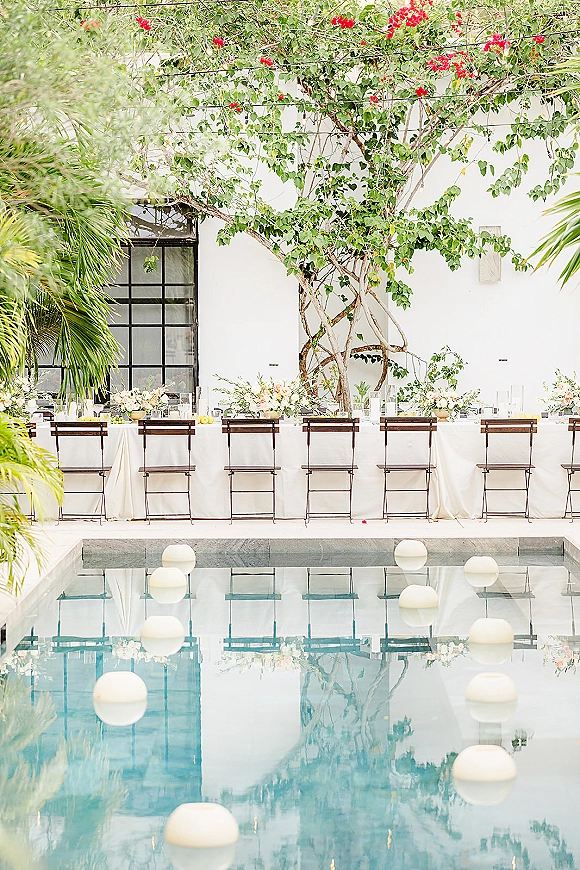 Reception tablescape with long banquet table setup, white linens, folding chairs, bud vases and glass hurricane candles by a poolside stucco wall