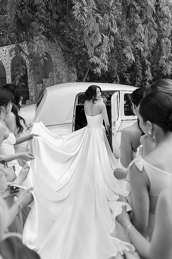 Bride getting ready as bridesmaids help arrange her long bridal gown train beside a vintage car on a tree-lined driveway by a stone building
