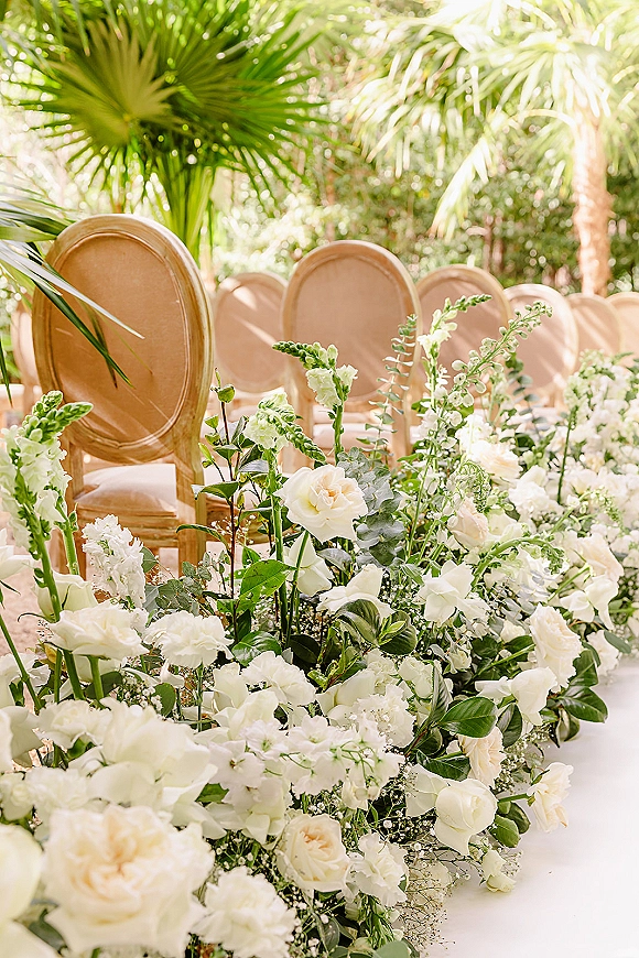 Ceremony aisle decor with wedding aisle flowers of white roses and greenery lining upholstered chairs in a sunlit tropical garden with palms