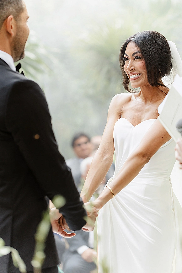 Wedding vows as bride and groom holding hands during an outdoor wedding ceremony, her veil flowing while guests and officiant look on