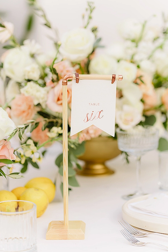 Wedding table number table number card in a brass holder on a copper stand, surrounded by blush florals, greenery, plates, goblet, lemons