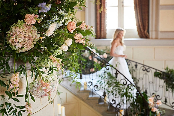 Wedding staircase decor with staircase floral installation of pastel blooms and greenery garland on wrought iron railing, candles by a window-lit stairway