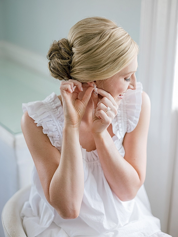 Bridal getting ready bride putting on earrings in a ruffled robe, showing engagement ring, blonde low bun, soft window light by a chair