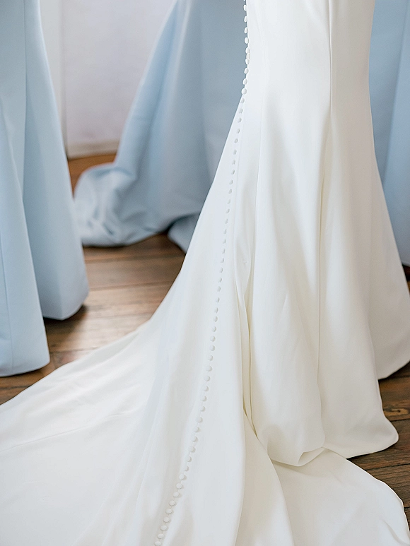 Wedding dress detail with covered button wedding dress back and satin train draped on a wood floor beside light blue curtains