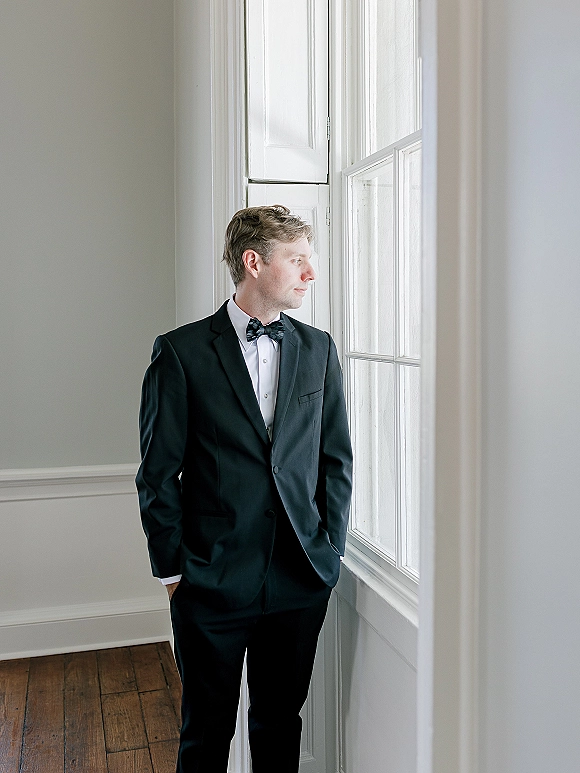 Groom portrait in a black tuxedo with bow tie, hands in pockets by a large window, lit by natural light against paneled wall