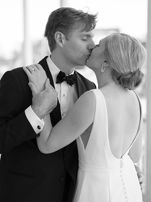 Wedding kiss portrait of bride and groom kissing in window light, groom in black tuxedo and bow tie, bride in backless gown with hoop earrings