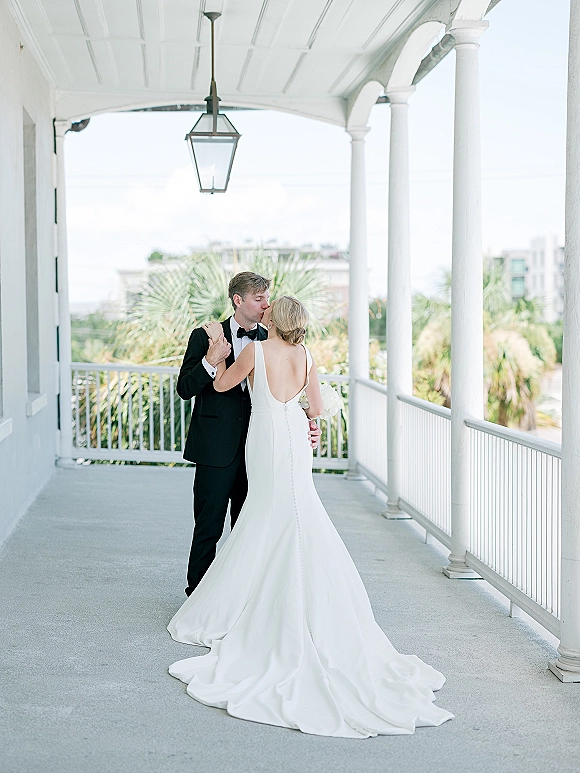 Wedding couple portrait of bride and groom kissing on a white porch with columns, her satin dress train and bouquet in soft lantern light