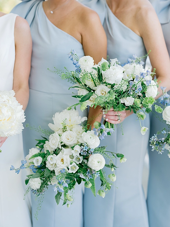 Bridesmaid bouquets of white and blue bouquet blooms with greenery, held by women in light blue one-shoulder dresses in soft outdoor light