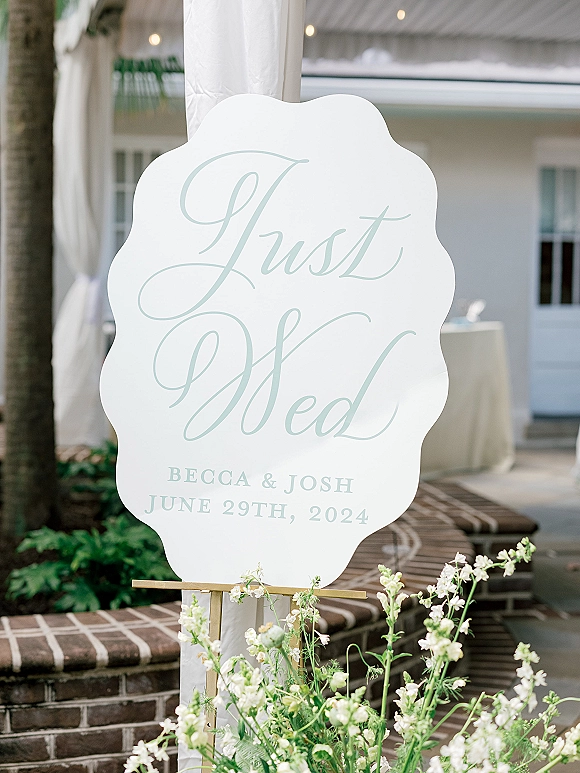 Wedding signage with calligraphy lettering on a gold stand, framed by white flowers and greenery on brick steps under a tent canopy