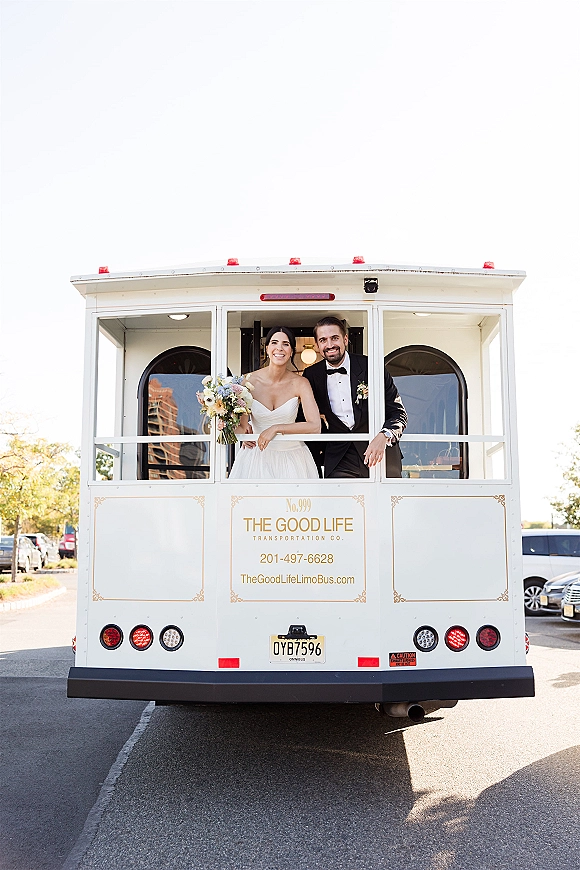 Couple portrait of bride and groom on trolley, leaning out the limousine bus window as she holds a wedding bouquet in a parking lot