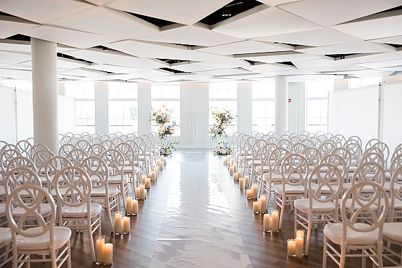 Ceremony setup for an indoor wedding ceremony with a white aisle runner, candle-lined aisle, clear acrylic podium, and bright windows backdrop