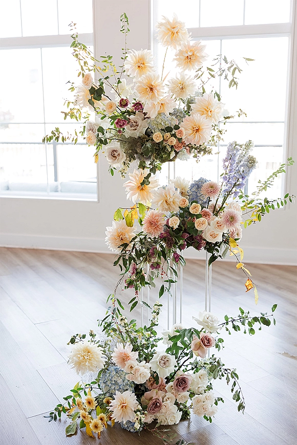 Wedding floral arrangement with dahlias and roses on ceremony floral pillars, set by large windows in a bright white room with wood floors