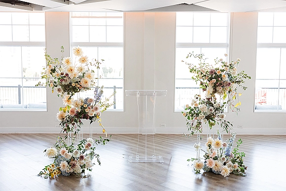 Ceremony altar decor with asymmetrical wedding arch flowers on clear acrylic stands, pastel roses and greenery in a bright room with large windows