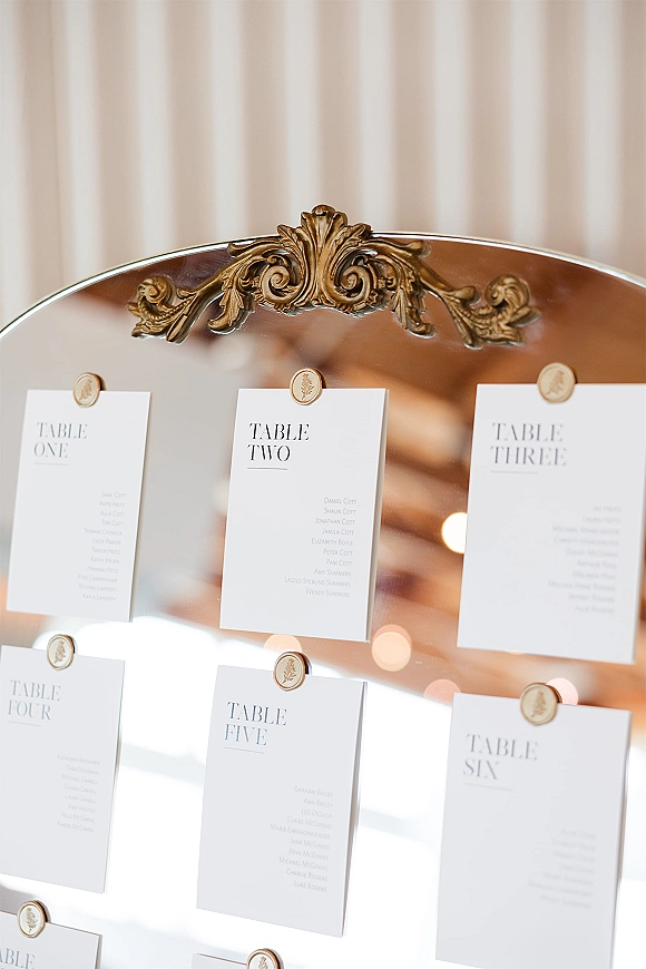 Wedding seating chart mirror display in an ornate gold frame with wax-sealed seating cards, set against a striped wall and warm bokeh lights