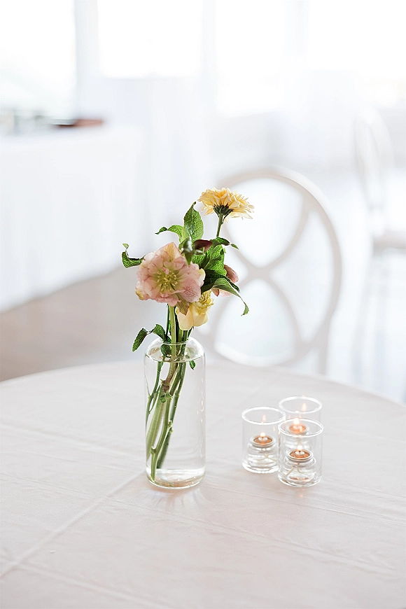 Wedding centerpiece with pastel flowers and greenery in a clear glass vase, surrounded by tea light candles on a white tablecloth in a bright reception room