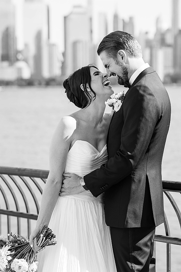 Couple portrait in a black and white wedding portrait, bride and groom laughing forehead-to-forehead by a waterfront railing with city skyline behind