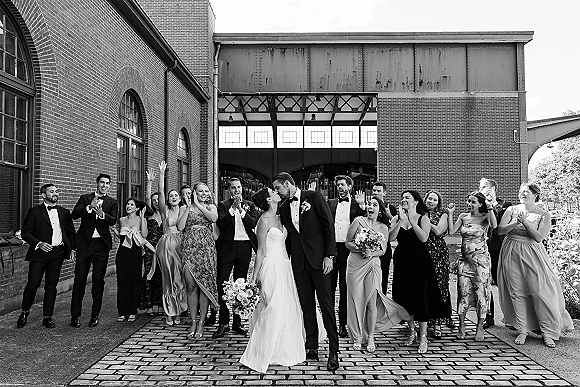 Wedding party photo of the bride and groom kiss as friends cheer, with bouquets and tuxedos on a cobblestone street by brick windows