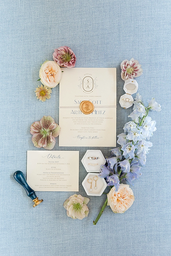 Invitation suite wedding invitation flat lay with gold wax seal, rings, pearl drop earrings, florals, and envelope liner on blue linen fabric
