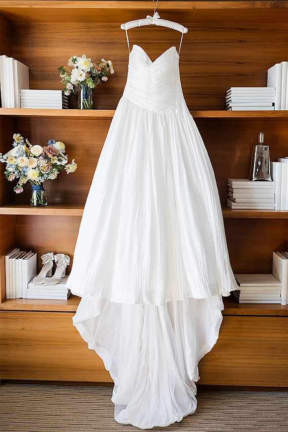 Wedding dress, strapless wedding dress with sweetheart neckline on a padded hanger amid florals, shoes, and books on wooden shelves