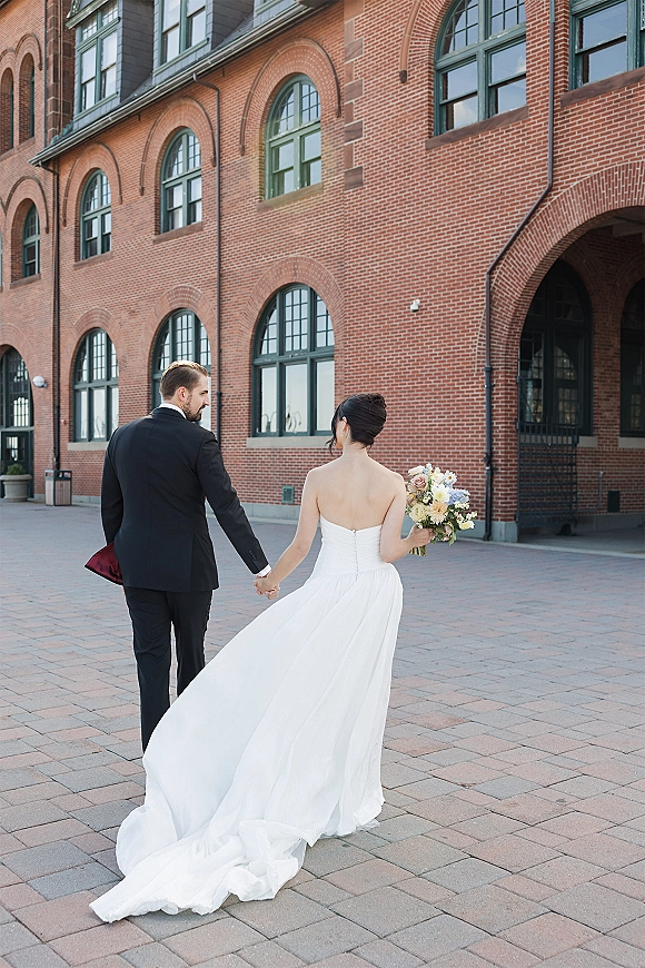 Couple portrait of newlyweds holding hands, walking away in a courtyard by a brick building with arched windows, bride in strapless gown with long train