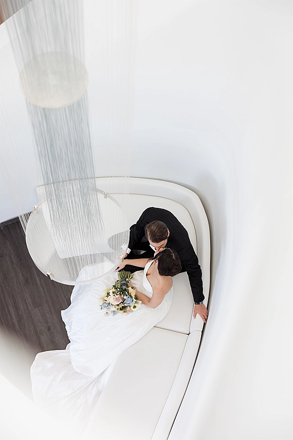Wedding kiss portrait of bride and groom kissing overhead on a white sofa, bride holding a pastel bouquet in a clean white room