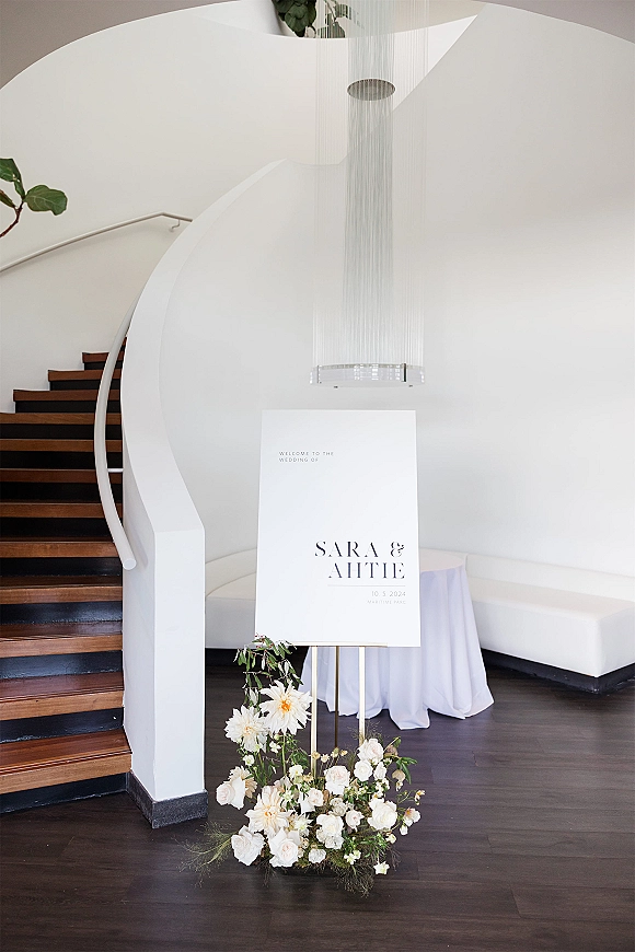 Wedding welcome sign on an easel with white roses and daisies, set by a curved staircase in a bright foyer with chandelier