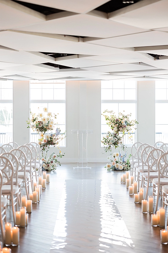 Ceremony setup for an indoor wedding ceremony with a white aisle runner, pillar candles, and florals by large windows with waterfront view