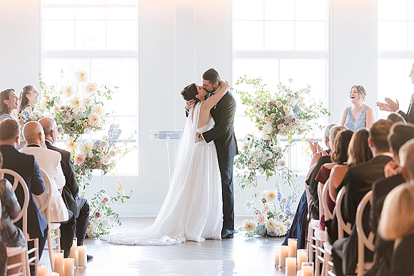 Wedding kiss as bride and groom embrace under a floral arch, long veil trailing by candle-lined aisle in bright indoor space with guests seated