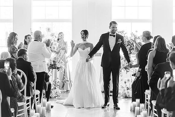 Wedding recessional as bride and groom walk the aisle holding hands, bride waving, guests cheering in bright indoor space with candles