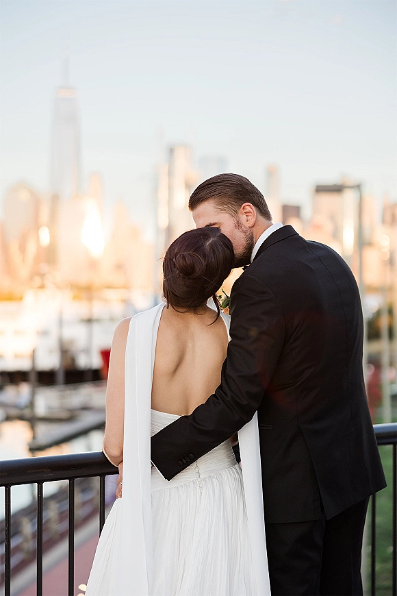 Wedding couple portrait of groom kissing bride’s forehead as they hug at a waterfront railing, her low back gown and cape facing the city skyline