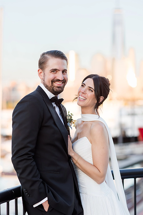 Couple portrait of bride laughing in a strapless dress with neck scarf beside groom in tuxedo at marina railing with skyline and boats