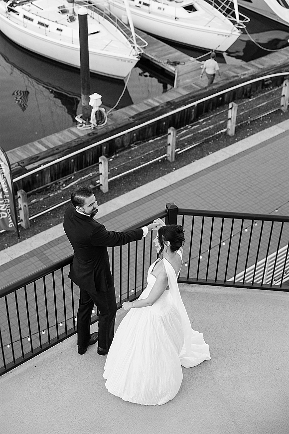 Couple portrait of bride and groom dancing as he twirls her in a flowing veil on a dock, marina sailboats behind and string lights overhead