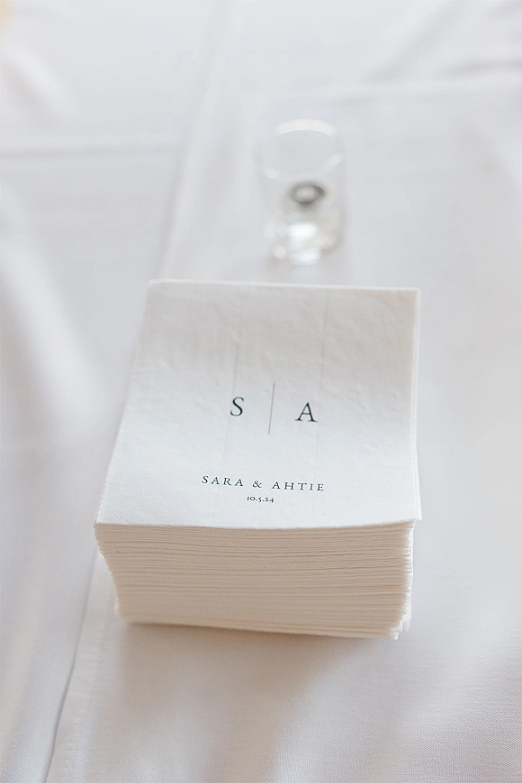 Wedding napkins with monogrammed cocktail napkins stacked beside wedding rings in a glass votive on a white tablecloth runner